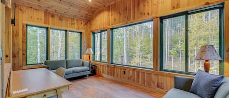 Interior de una cabaña con paredes de madera, ventanas amplias, un sofá, una mesa y una lámpara.