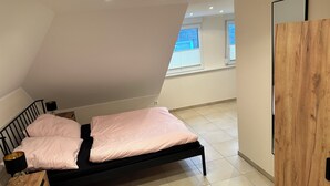 A bedroom with a black bed, a wooden nightstand, a small table, and a window with blinds. - Vacation apartment / trade fair apartment - quietly located on the outskirts of Krefeld  (Krefeld)