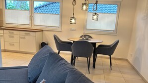 A modern living room with a sofa, dining table, and chairs. There are windows with blinds, a kitchen area, and hanging pendant lights. - Vacation apartment / trade fair apartment - quietly located on the outskirts of Krefeld  (Krefeld)
