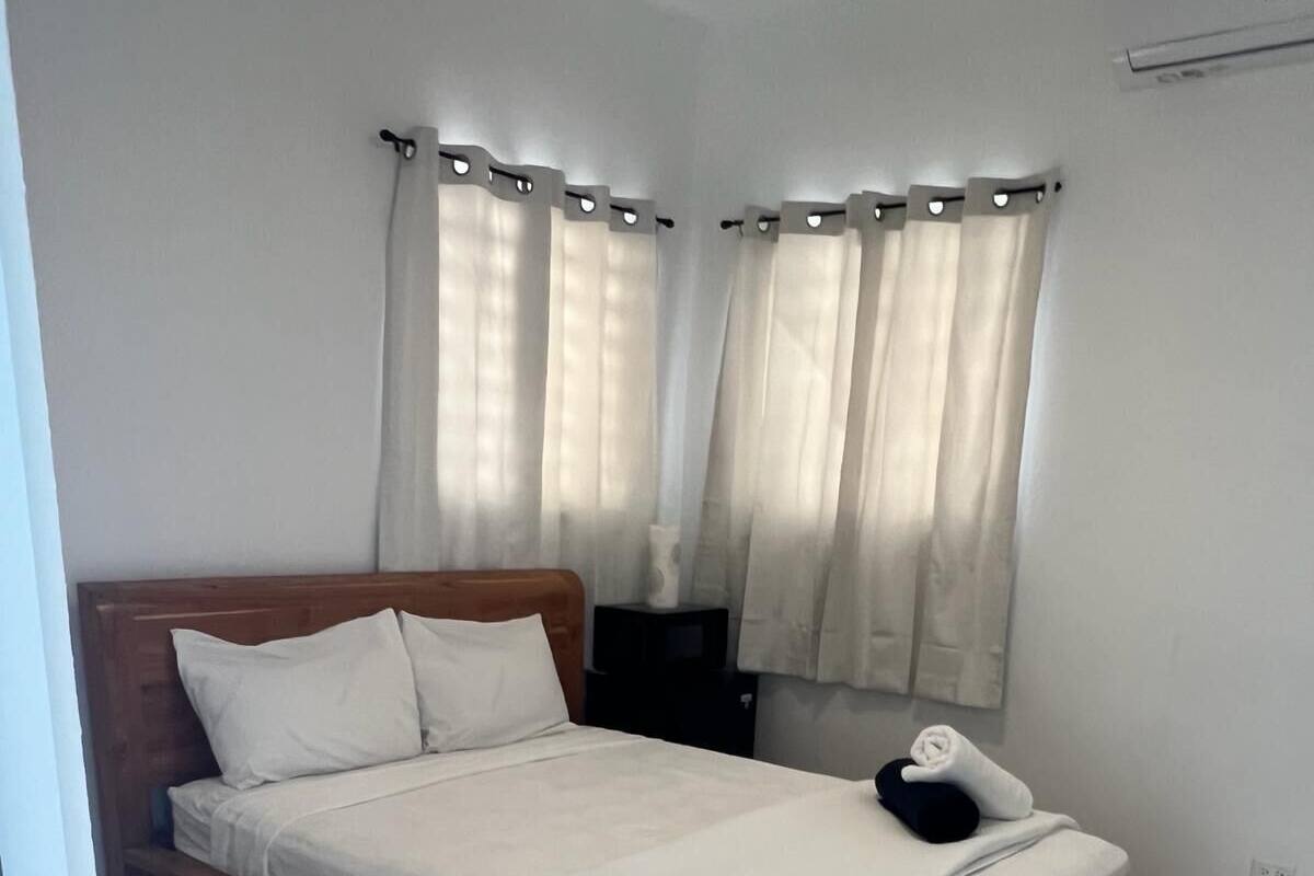 Ein ordentlich bezogenes Bett mit Holzkopfseitenverkleidung, einem Nachttisch und einem Fenster mit Vorhängen.