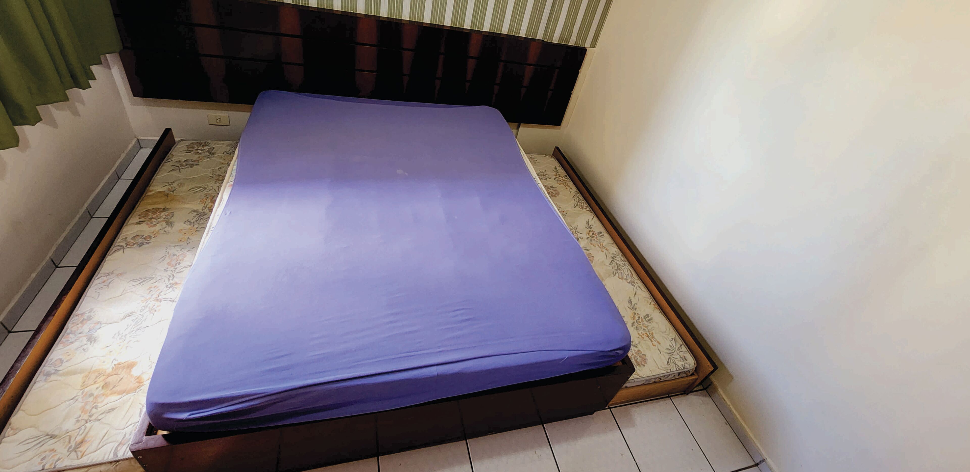 Un lit avec un matelas violet dans une pièce dont le sol est recouvert de carreaux et qui possède une fenêtre ornée de rideaux verts.