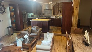 Amplia sala de estar con una mesa de centro de madera, sofás blancos y una mesa de comedor de madera. Se observa una cocina con armarios de madera oscura y encimera de granito al fondo.