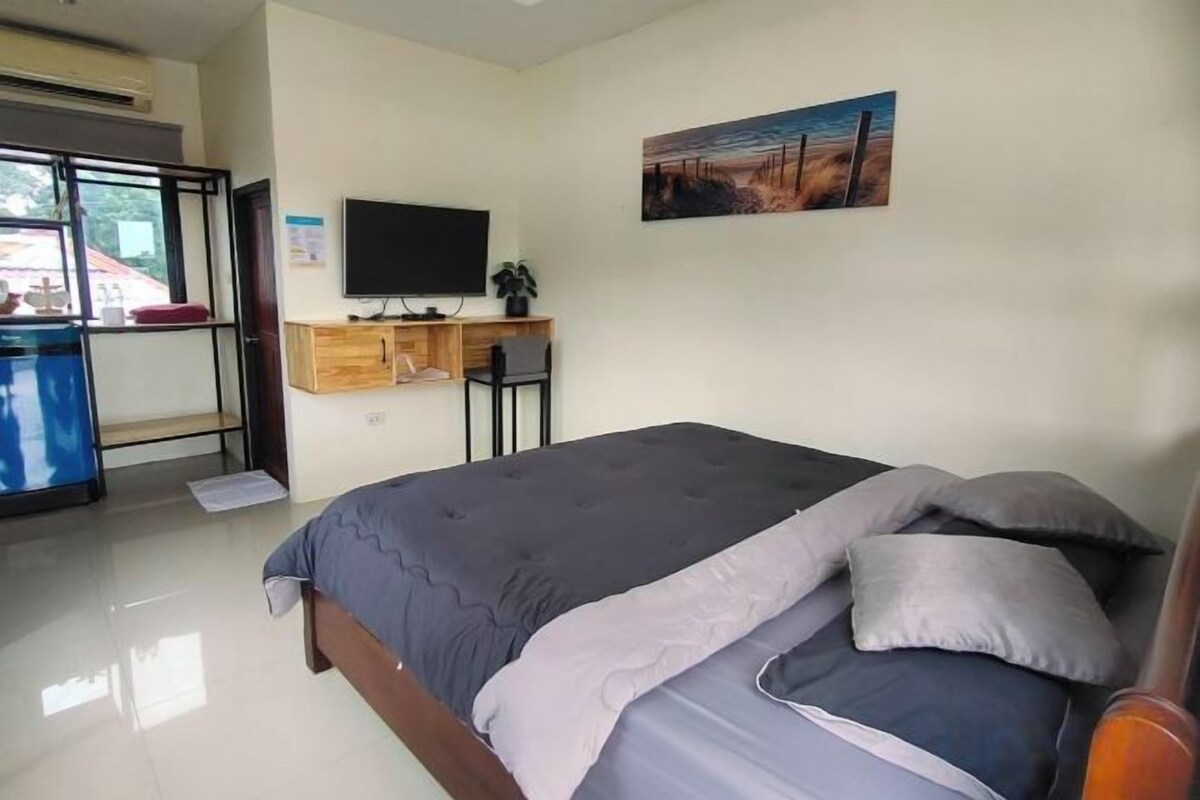 A bedroom with a bed, a desk, a chair, a television, and a window.