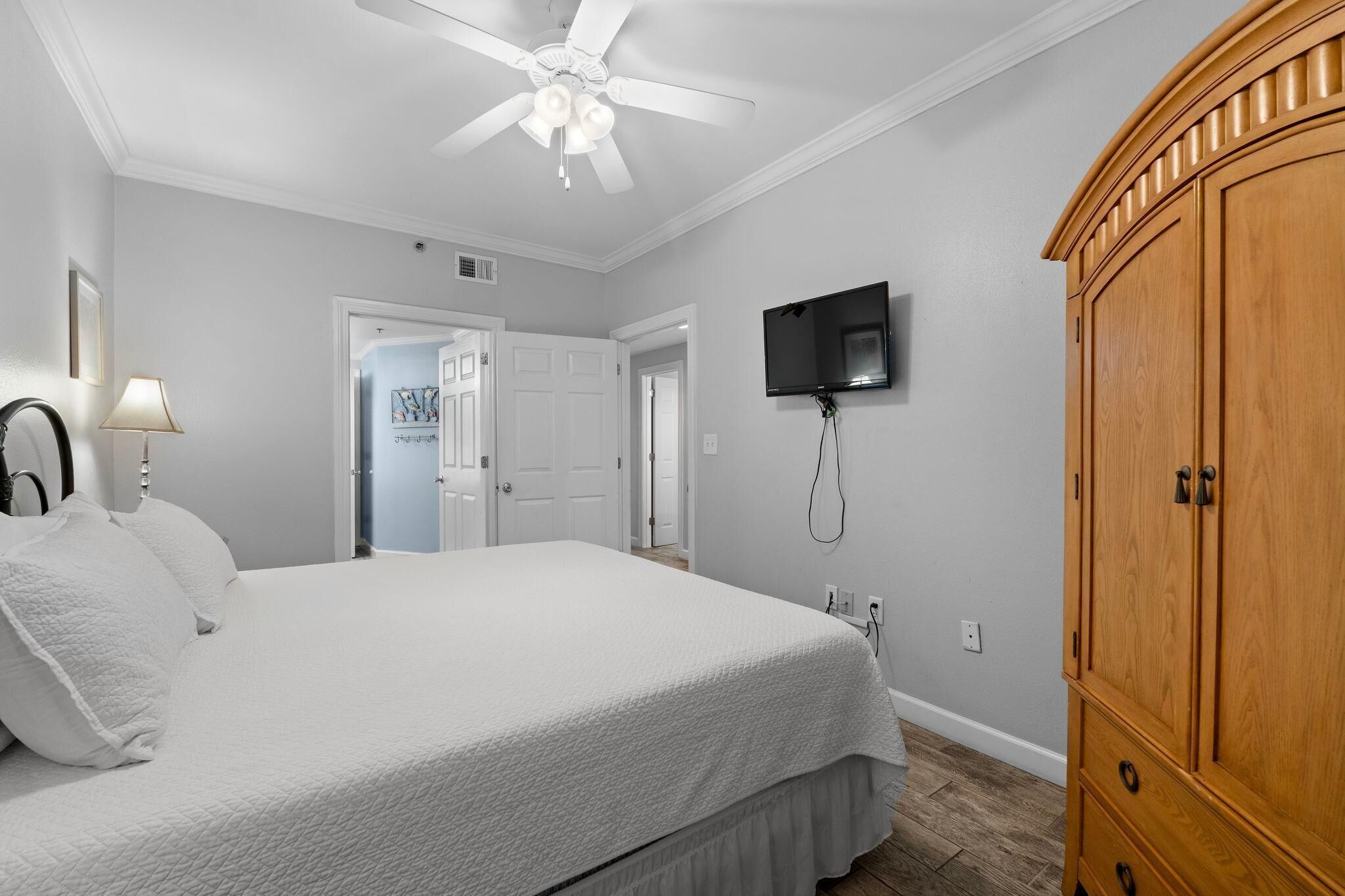 A bedroom with a bed, a ceiling fan, a TV mounted on the wall, and a wooden wardrobe.