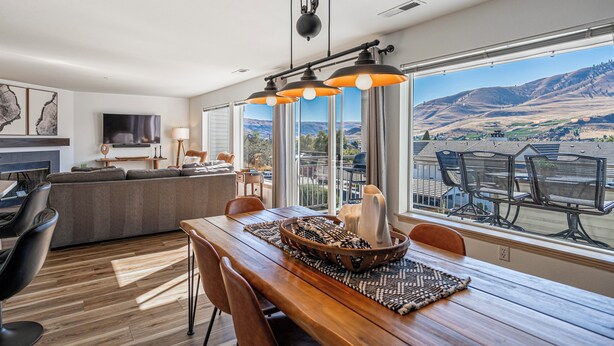 A modern living room with a wooden dining table, black chairs, and a large window offering a view of hills. - Chelan Condo | Breathtaking Mountain & Lake Views (Chelan)
