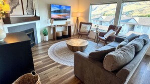 A modern living room with a fireplace, a large TV mounted on the wall, a sofa set, and a view of a mountainous landscape through large windows. - Chelan Condo | Breathtaking Mountain & Lake Views (Chelan)