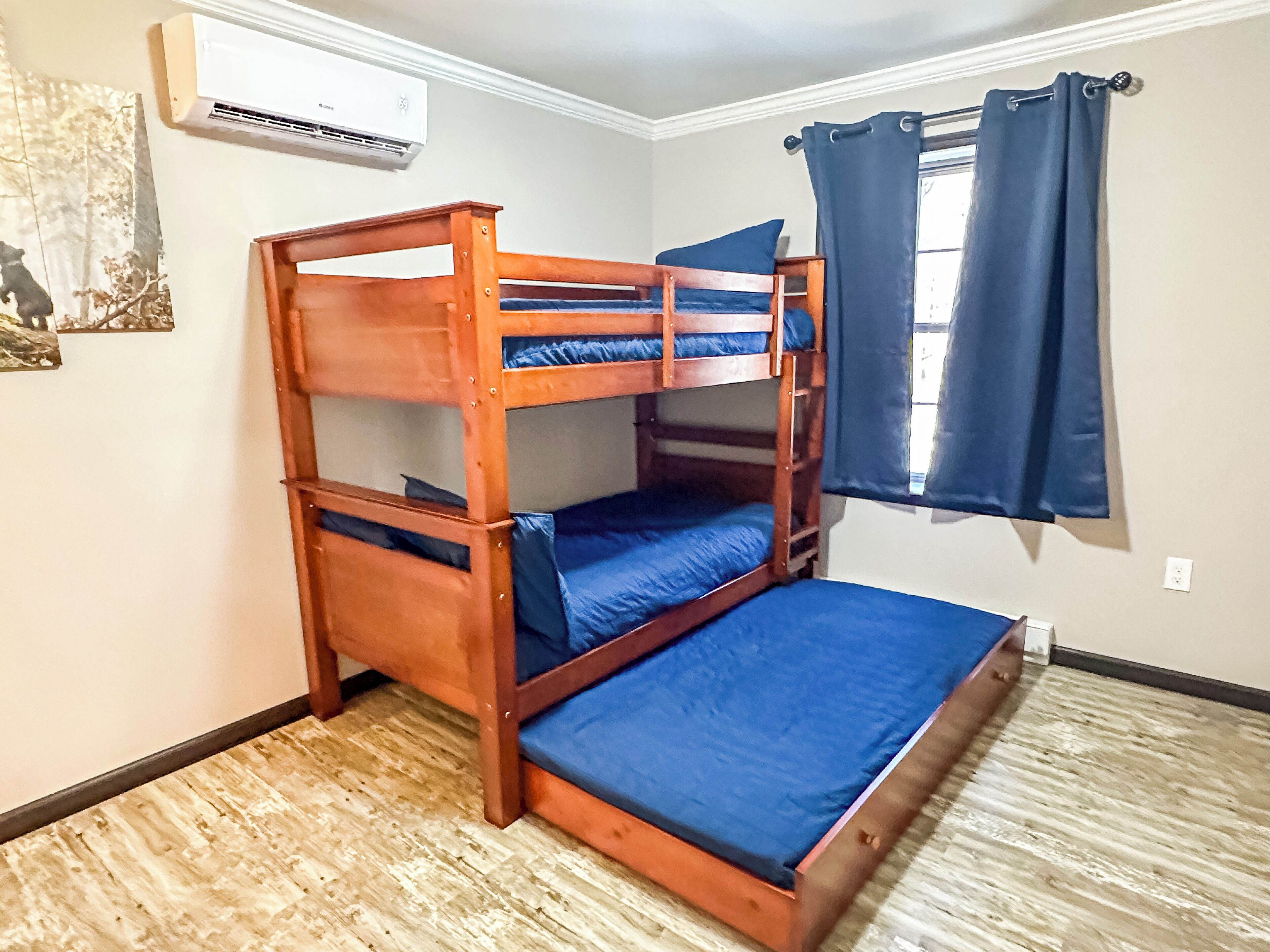 A bunk bed with blue bedding in a room with a wooden floor, an air conditioning unit, and a window with blue curtains.
