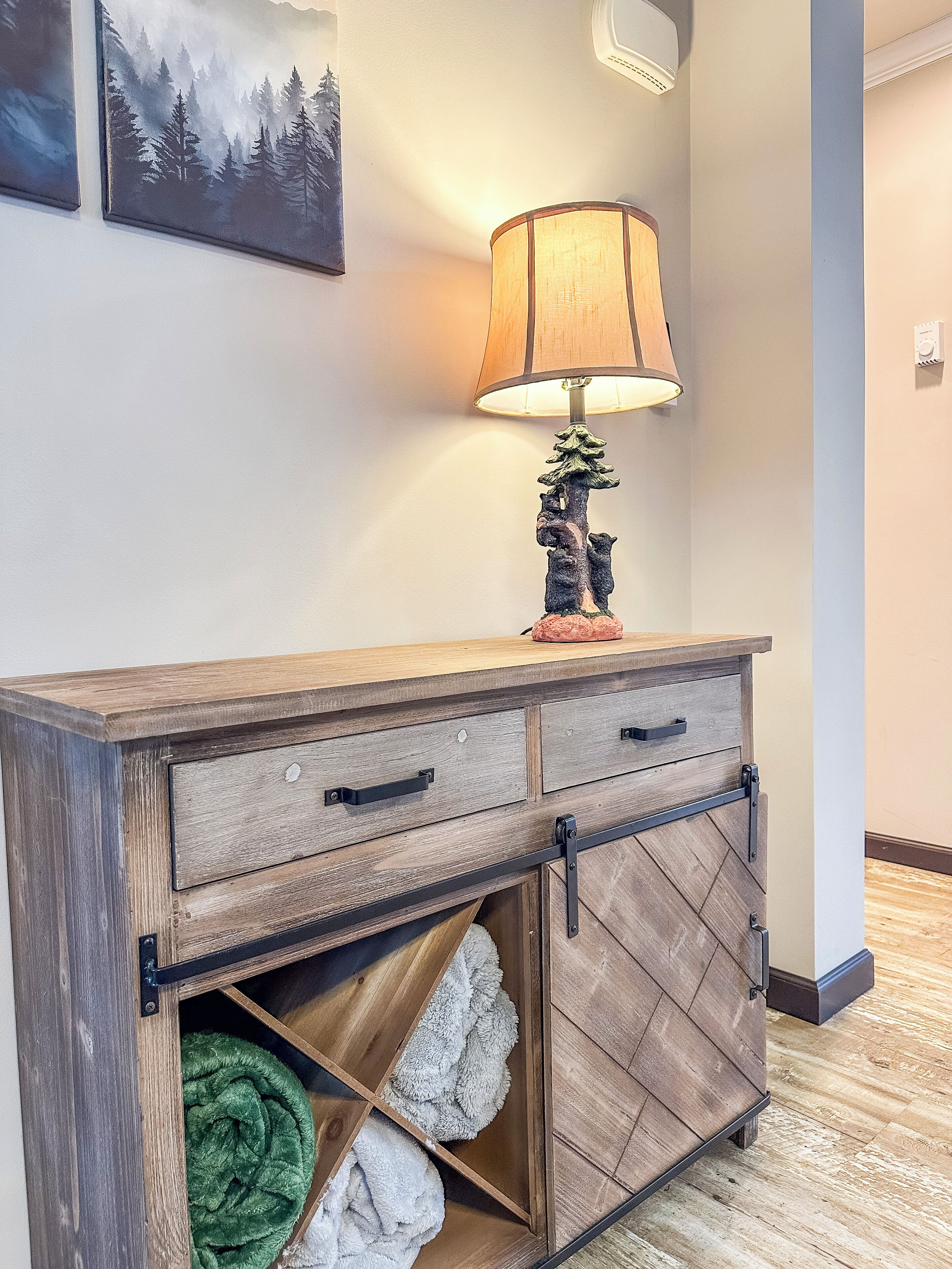 Een houten kastje met twee lades en een plank, een lamp met een ontwerp van een boom, en een groen-wit dekbed op de plank.