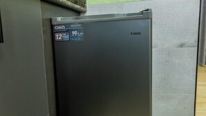A compact refrigerator with a digital display, situated in a corner of a room with a green textured wall and a grey floor. - Cozy Residences Kampala 008 (Kampala)