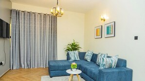 A living room with a blue sofa, a small white table with a vase, a potted plant, and a grey curtain. - Cozy Residences Kampala 008 (Kampala)