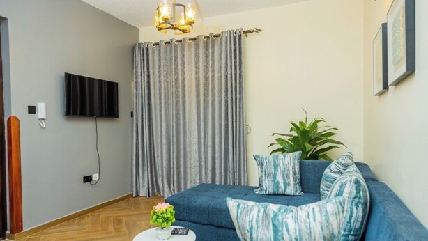 A living room with a blue sofa, a small round table with a plant, a TV mounted on the wall, and a curtain. - Cozy Residences Kampala 008 (Kampala)