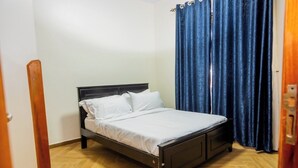 A single bed with a wooden frame and white bedding in a room with wooden flooring and a blue curtain. - Cozy Residences Kampala 008 (Kampala)