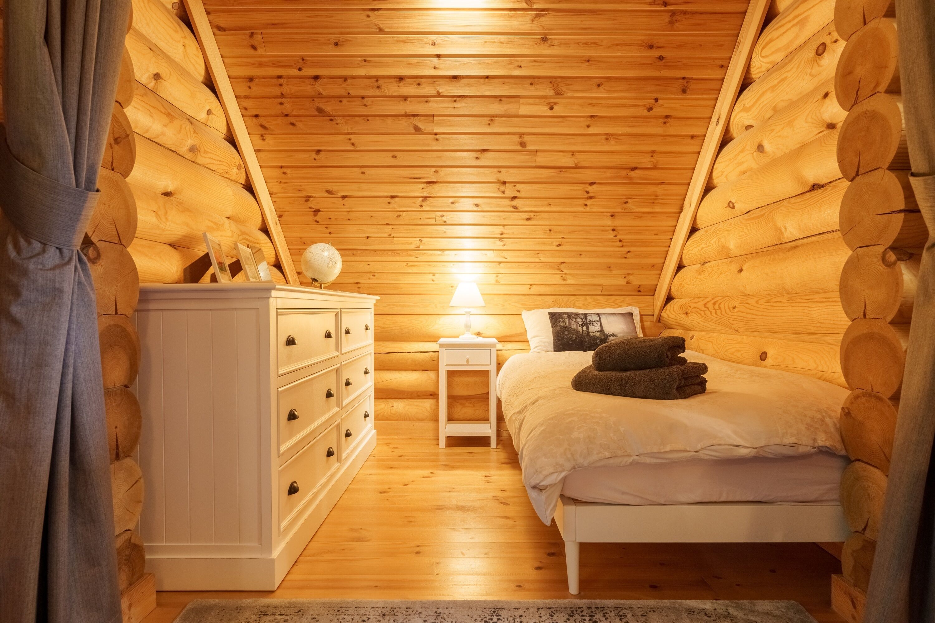 A cozy log cabin bedroom with a bed, dresser, lamp, and a globe on a nightstand.