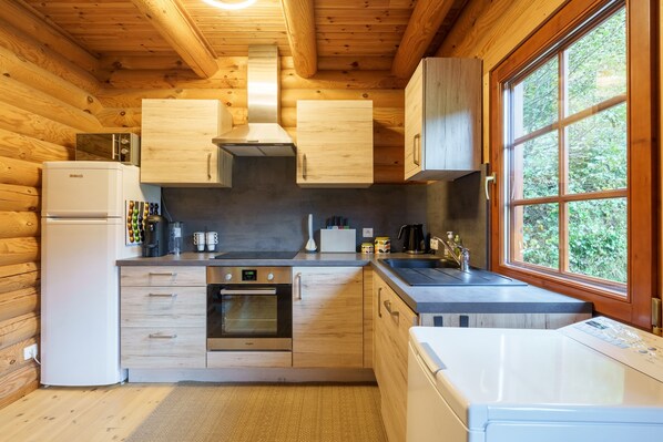 A compact kitchen with wooden walls, a refrigerator, an oven, a sink, and a window with a view of greenery.