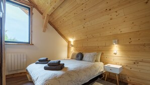 A bedroom with a sloped wooden ceiling, a large window, a bed with a floral bedspread, two pillows, a bedside table with a lamp, and a radiator.