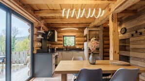 A modern log cabin interior with a wooden dining table, two chairs, a vase with flowers, and a kitchen area with a large window.