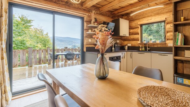 A wooden cabin interior with a dining area, a kitchen, and a large window overlooking a deck.