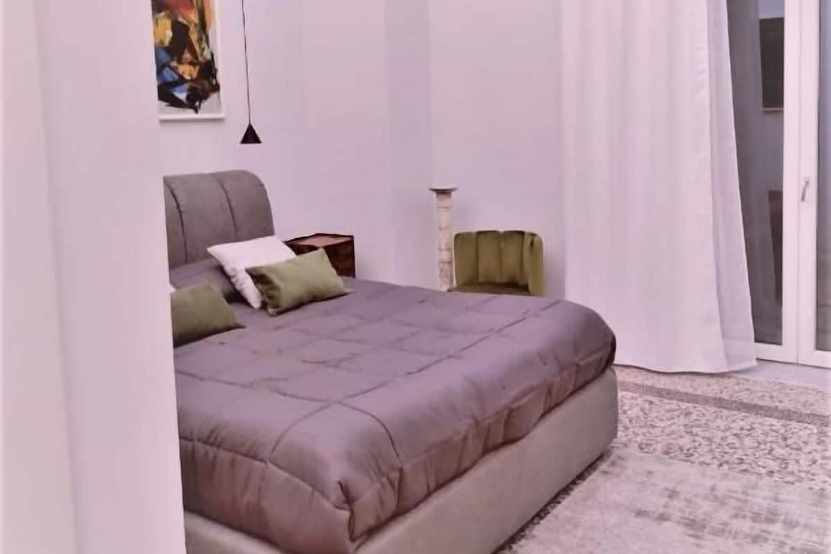 Una camera da letto con un letto, un comodino, una lampada, un quadro e vista sull'esterno.