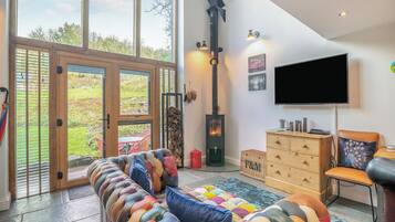 A spacious living room with a large window, a fireplace, a sofa with colorful cushions, a television, and a wooden cabinet.