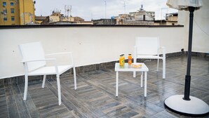 A rooftop terrace with two white chairs, a small table with two orange juice bottles, and a floor lamp.