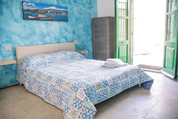 Una cama con una colcha estampada, una mesita de noche, un armario alto y una puerta que da al exterior.