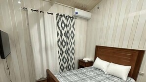 Ein Schlafzimmer mit einem hölzernen Bett, einer grau-weißen karierten Tagesdecke, einem grauen Teppich, Holzboden und einem weißen Vorhang mit einem schwarzen Rautenmuster. Ein Deckenventilator ist ebenfalls vorhanden.