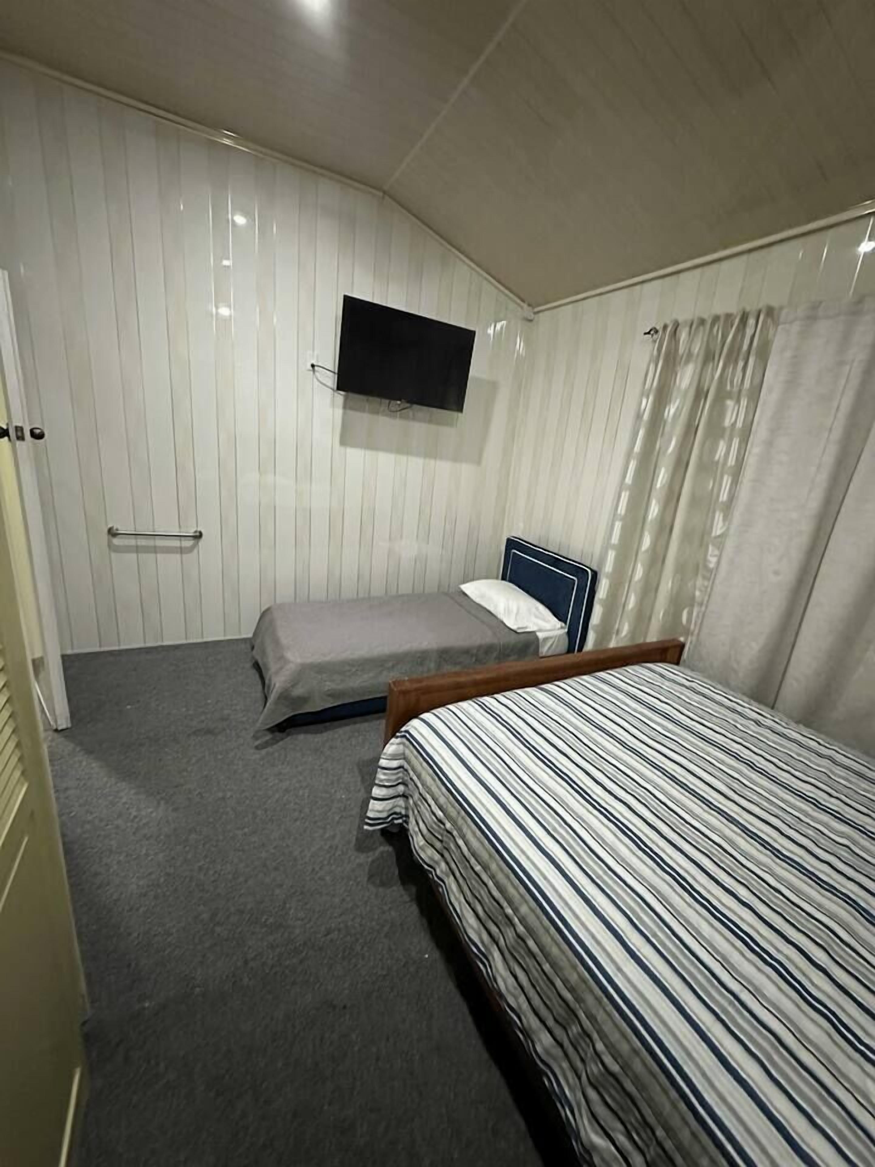 Een kleine, compacte kamer met een eenpersoonsbed, een nachtkastje, een aan de muur bevestigde televisie en een gordijn voor het raam.