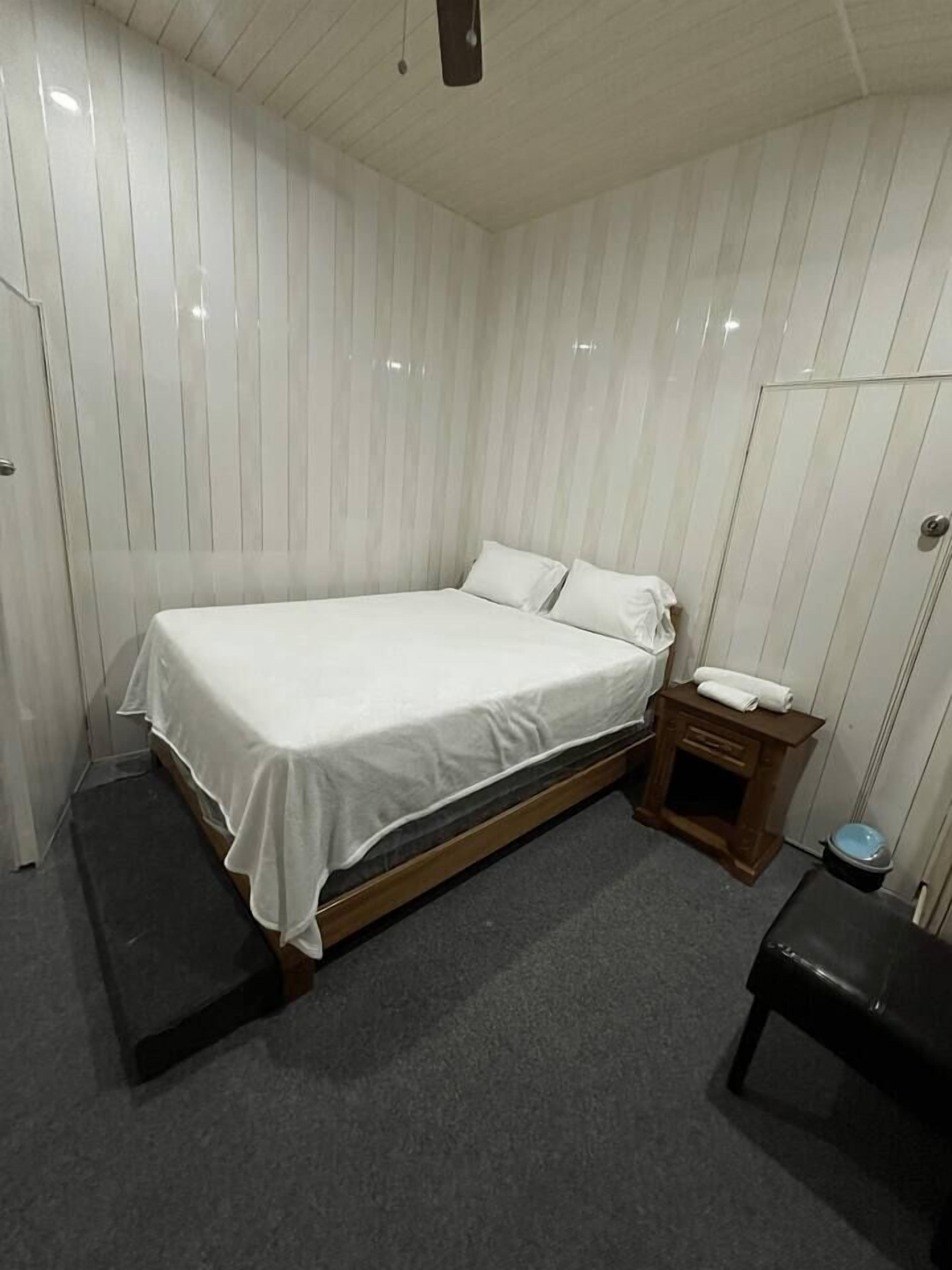 A single bed with a wooden frame and white bedding in a room with white paneled walls and a grey carpet.