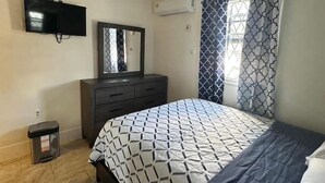 Un dormitori amb llit, ventilador de sostre, televisió, prestatge i finestra amb cortines estampades.