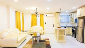 Una sala de estar moderna con dos sofás blancos, una mesa de centro de vidrio y un tapete colorido. Junto a ella, una cocina con refrigerador, estufa y armarios.