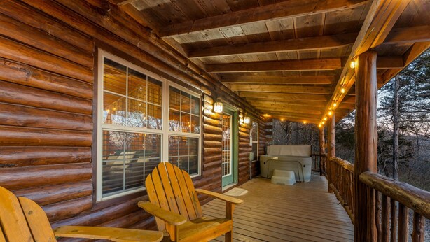 Une cabane en rondins avec une terrasse en bois, deux fauteuils Adirondack, un jacuzzi et une vue sur la forêt environnante.