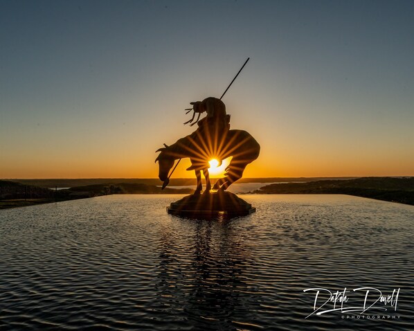 A statue of a figure with a spear, positioned on a platform in a body of water, with the sun setting in the background.