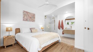 Un dormitorio moderno con una cama grande, lámparas de noche, ventilador de techo y baño con ducha, lavamanos y toallas.