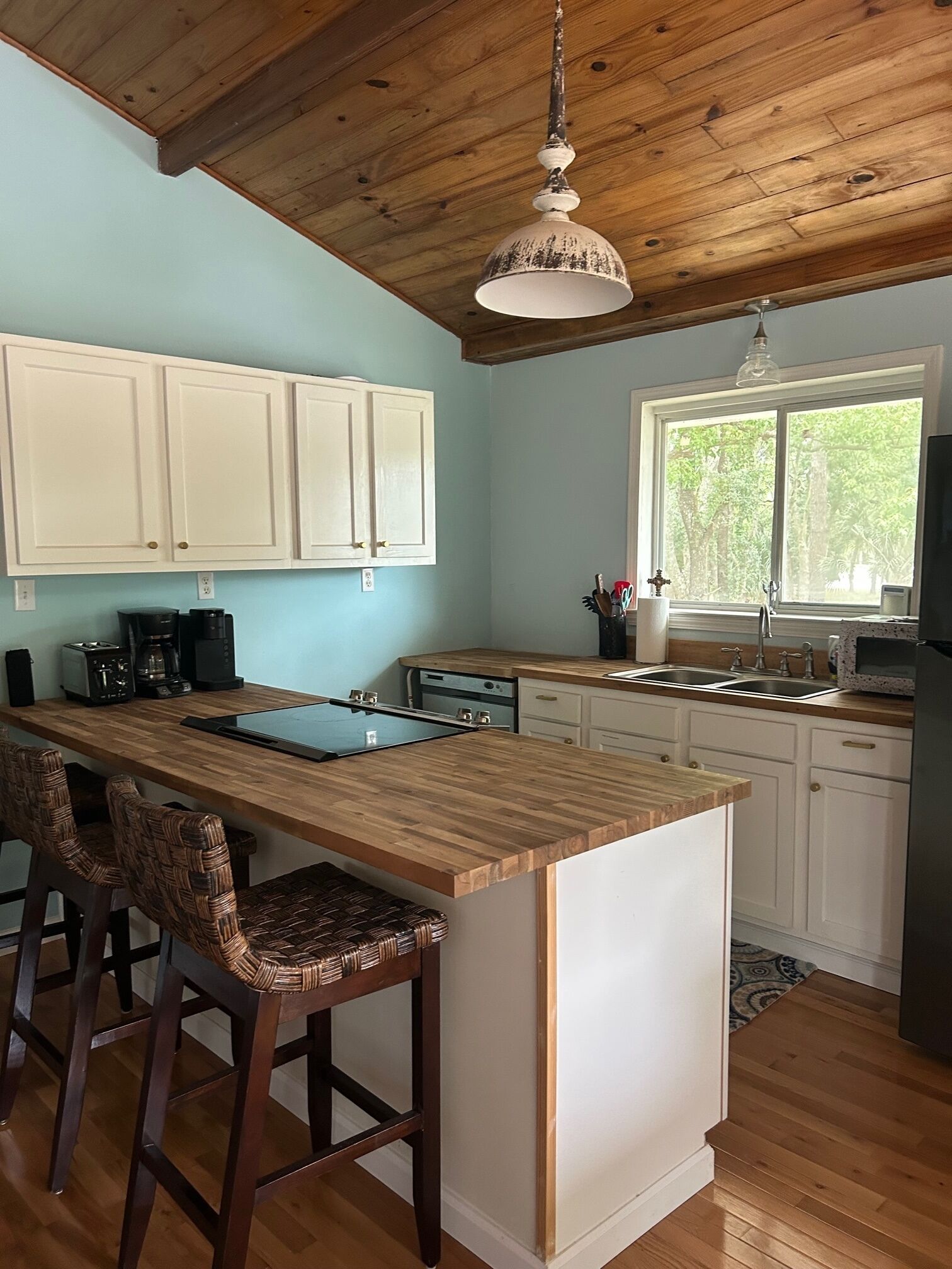 A kitchen with wooden cabinets, a wooden island with bar stools, a window with a view of greenery, and a hanging light fixture.