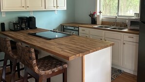 A kitchen with wooden cabinets, a wooden island with bar stools, a window with a view of greenery, and a hanging light fixture.