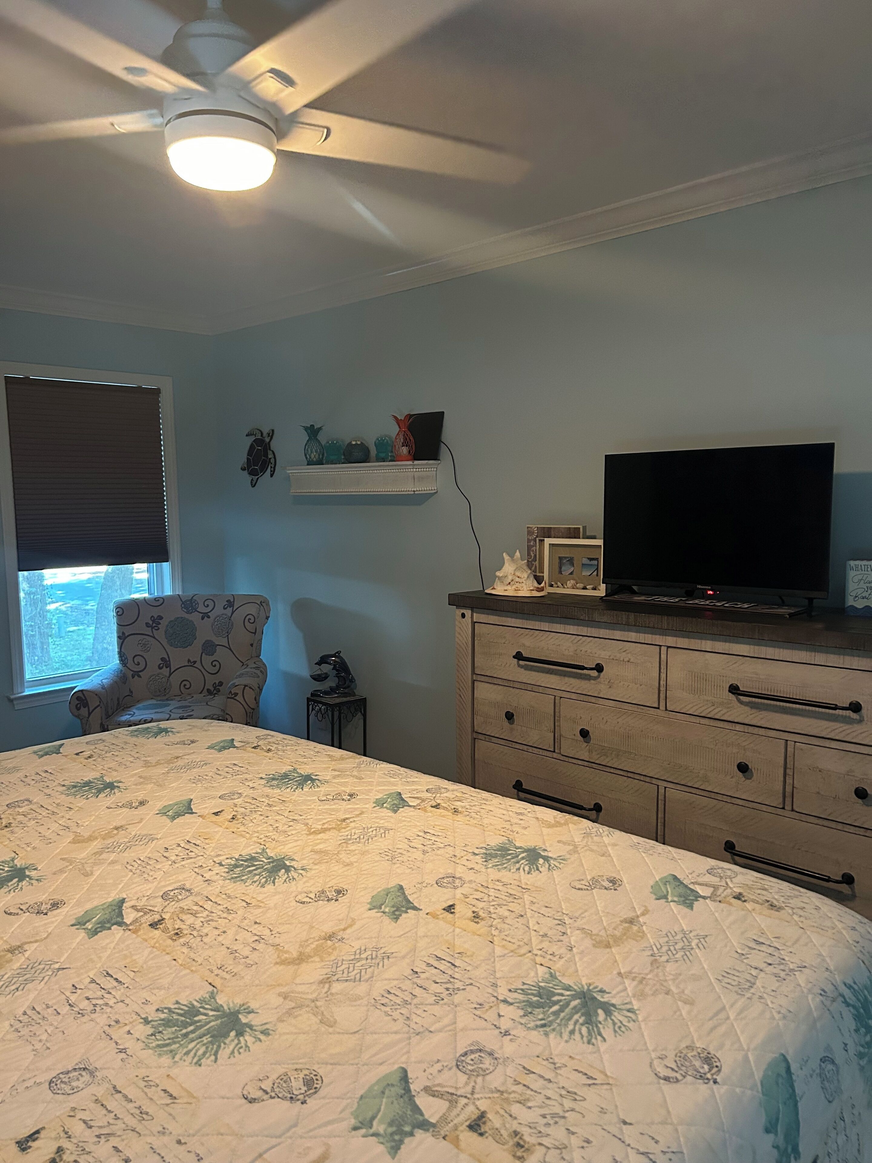 Une chambre à coucher comprenant un lit, une chaise, une commode, un téléviseur et un ventilateur de plafond.