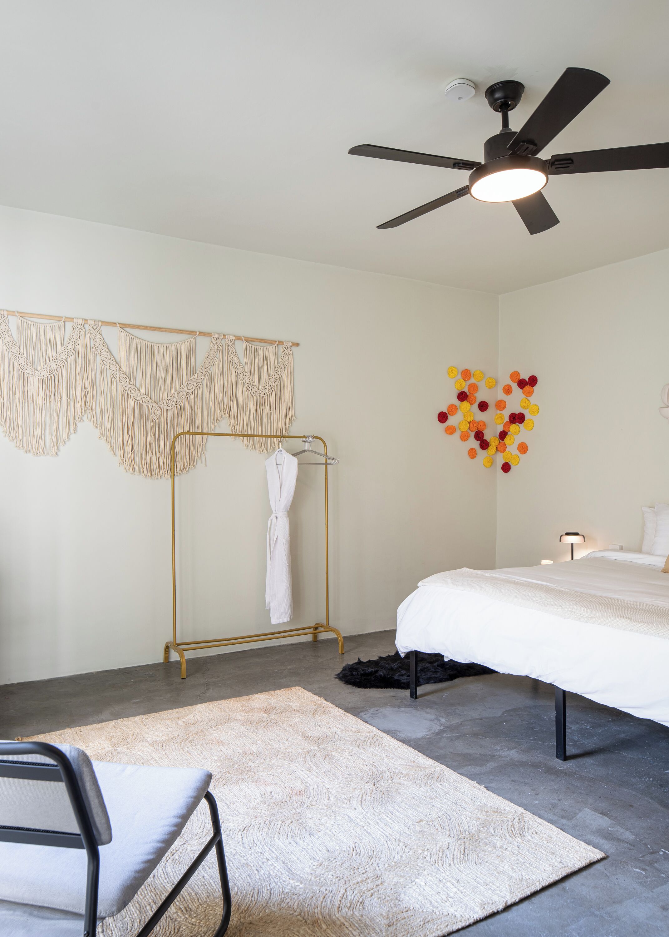 A modern bedroom with a bed, a ceiling fan, a macramé wall hanging, a gold-colored clothing rack, and a heart-shaped wall decoration.