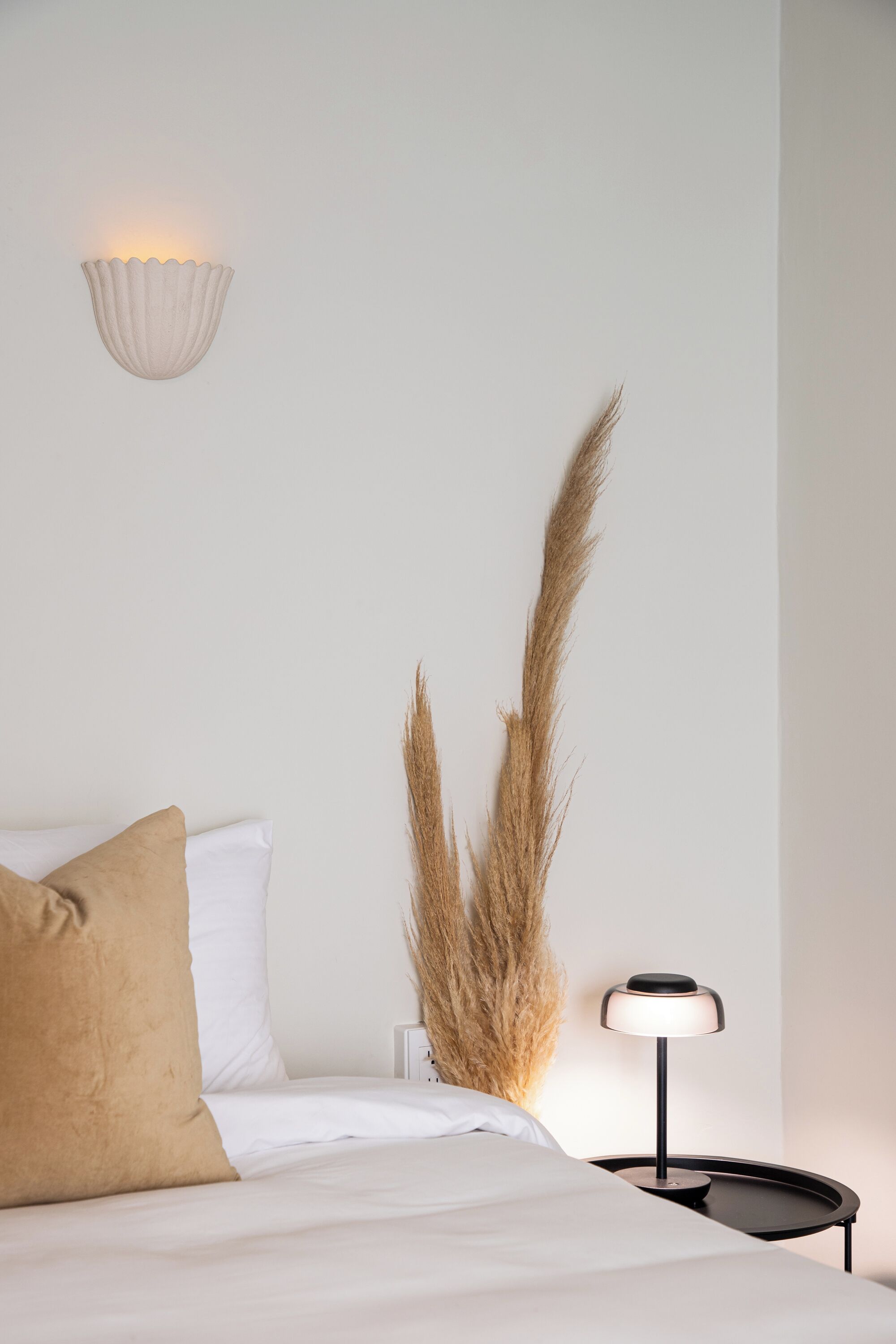 A bedroom with a bed, a bedside lamp, a wall-mounted light fixture, and pampas grass.