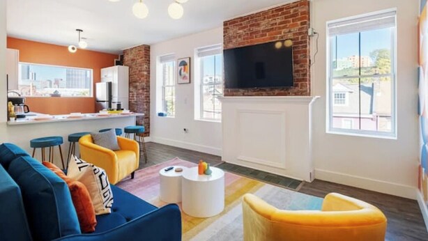 A modern living room with a brick accent wall, a flat-screen TV, and a city view. - Modern 1BR Brick+Kingbed+Free Parking+Close to DT (Pittsburgh)