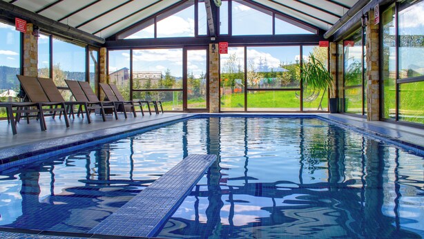 An indoor swimming pool with clear glass walls, lounge chairs, and a view of the outdoors. - Bucovina Residence & SPA (Gura Humorului)