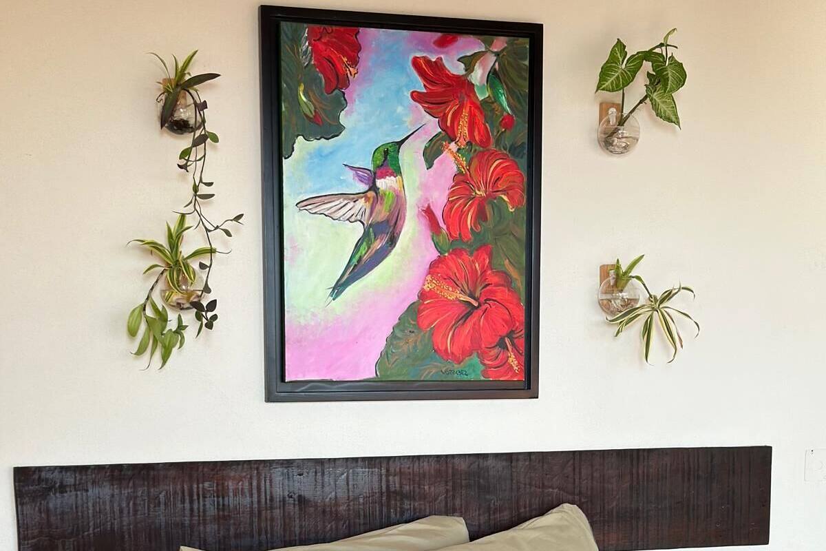 A bed with a colorful blanket, a dark wooden headboard, and a framed painting of a hummingbird and flowers on the wall.
