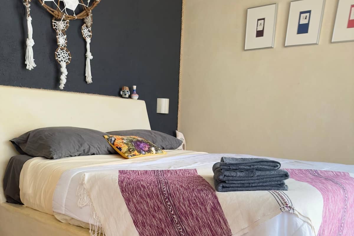 A bed with a white and purple blanket, a folded gray blanket, and a floral pillow.