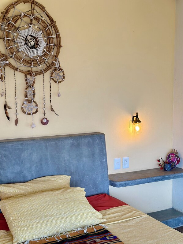 Ein Schlafzimmer mit Bett, Kissen, einem an der Wand befestigten Traumfänger, einer Wandleuchte und einem Regal mit Dekoration.