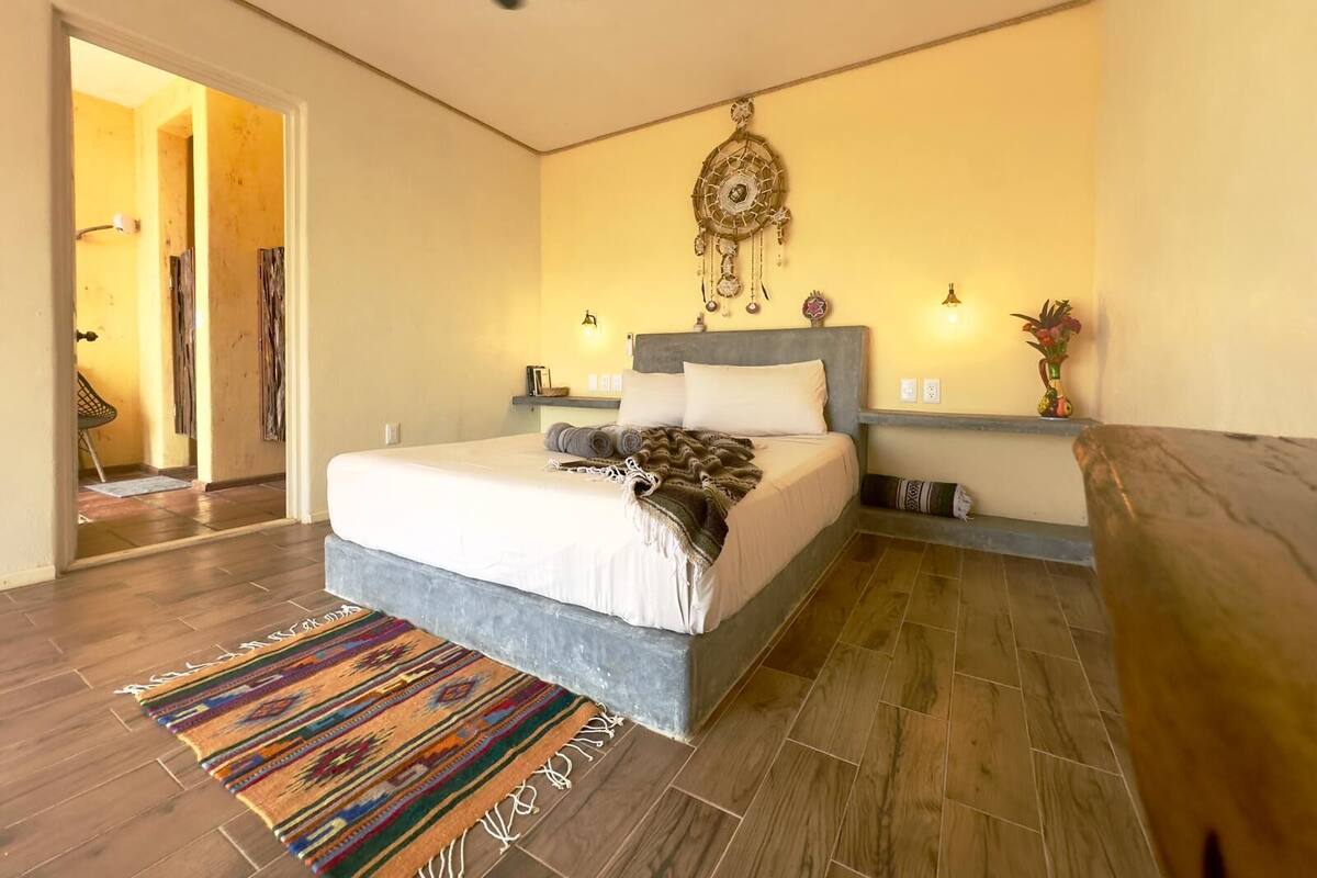 A bedroom with a bed, a ceiling fan, a wall-mounted dreamcatcher, and a small shelf with decorative items.