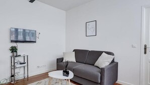 Una sala de estar moderna con un sofá gris, una pequeña mesa de centro blanca, un televisor montado y un cuadro enmarcado en la pared.
