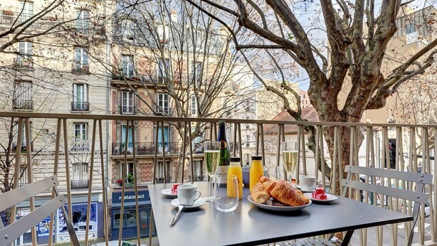 Mesa en el balcón con alimentos para el desayuno, un croissant y bebidas, con vistas a una calle y edificios de la ciudad.