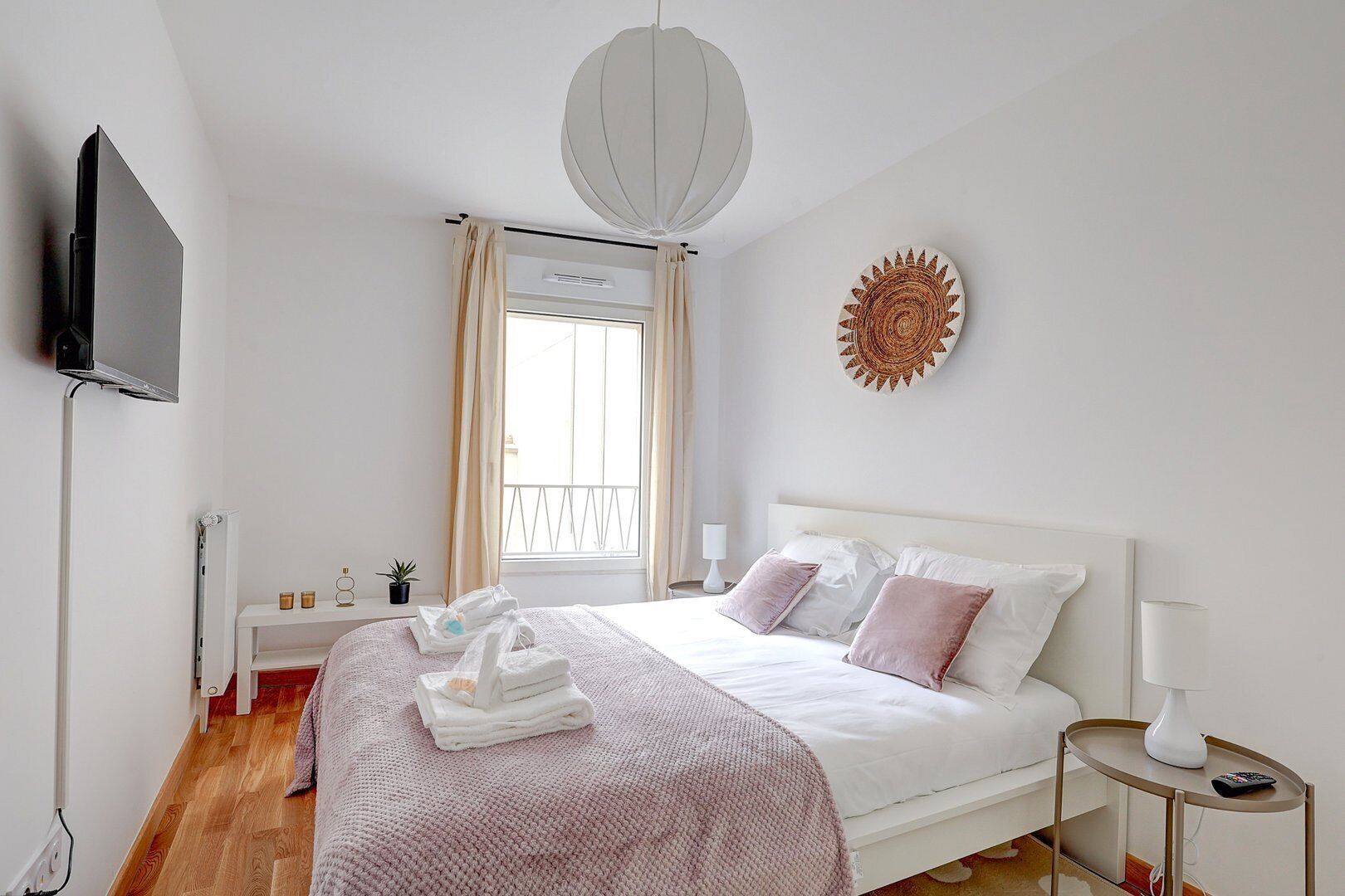 A bedroom with a bed, a TV mounted on the wall, a window with curtains, a wall-mounted artwork, and a bedside table with a lamp.