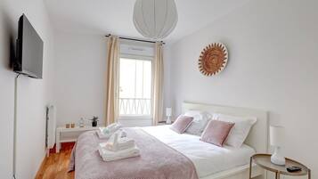 A bedroom with a bed, a TV mounted on the wall, a window with curtains, a wall-mounted artwork, and a bedside table with a lamp.