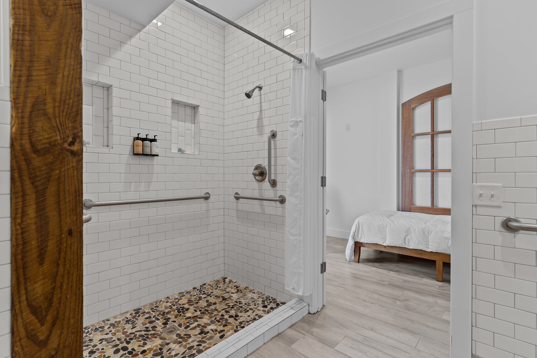 A modern bathroom with white subway tiles, a shower with a glass door, a wooden door frame, and a bed with a white bedspread in the adjacent room.
