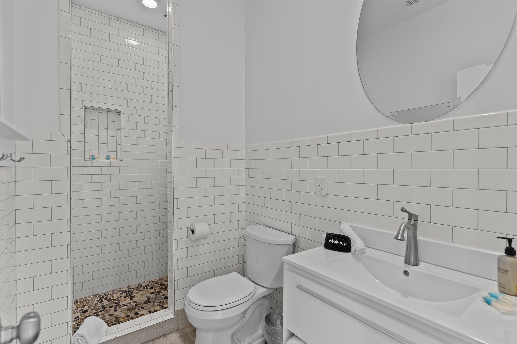 Une salle de bain moderne avec des carreaux de céramique blancs rectangulaires, une douche en verre, des toilettes et un meuble-lavabo avec un évier et un miroir.